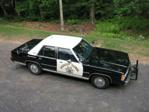 1991 Ford LTD Crown Victoria - Elevated view of a black-and-white police cruiser marked Highway Patrol with a gold star door shield and front push bumper, parked beside a…