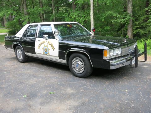 1991 Ford LTD Crown Victoria - Front three-quarter view of a black-and-white police sedan with twin pillar spotlights, chrome grille, and Highway Patrol door markings.