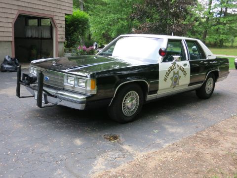 1991 Ford LTD Crown Victoria - Front three-quarter view of a black-and-white Highway Patrol cruiser with a front push bumper, parked near an open garage and shrubs.