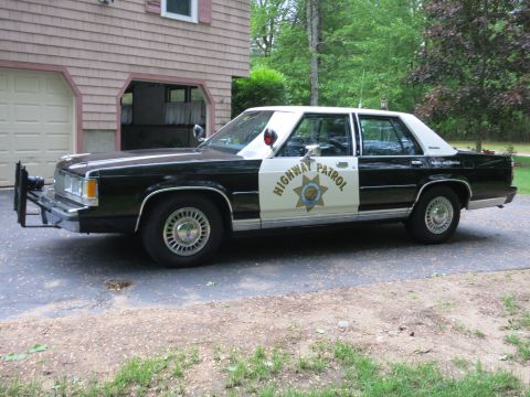 1991 Ford LTD Crown Victoria - Driver-side front three-quarter view of a black-and-white Highway Patrol cruiser with a push bumper, parked beside a shingled house.