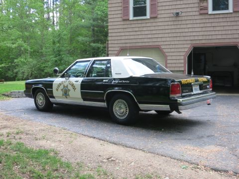 1991 Ford LTD Crown Victoria - Driver-side rear three-quarter view of a black-and-white Highway Patrol cruiser with a gold star door shield, parked beside a house.