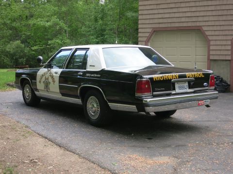 1991 Ford LTD Crown Victoria - Driver-side rear three-quarter view of a black-and-white police sedan with yellow Highway Patrol trunk lettering, parked near a garage.