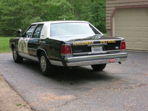 1991 Ford LTD Crown Victoria - Driver-side rear three-quarter view of a black-and-white police sedan with yellow Highway Patrol trunk lettering, parked by a garage.