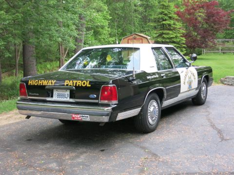 1991 Ford LTD Crown Victoria - Passenger-side rear three-quarter view of a black-and-white Highway Patrol cruiser with yellow trunk lettering, parked near trees.