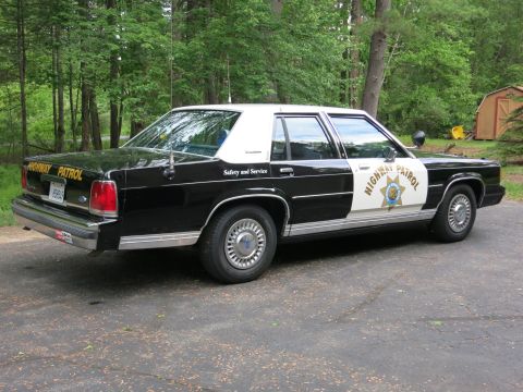 1991 Ford LTD Crown Victoria - Passenger-side rear three-quarter view of a black-and-white Highway Patrol cruiser with a gold star door shield, parked by woods.