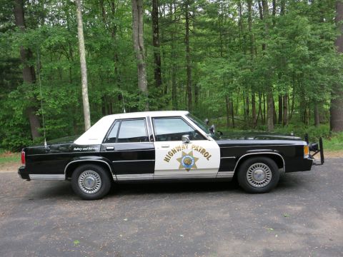1991 Ford LTD Crown Victoria - black-and-white Highway Patrol police sedan with a gold star emblem and tall whip antenna parked on pavement at the edge of green woods.