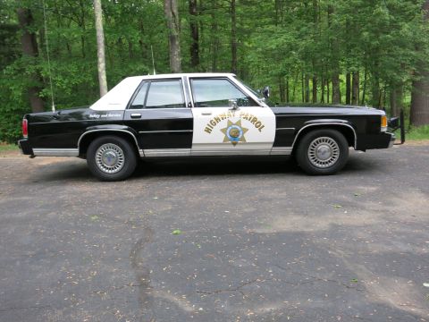 1991 Ford LTD Crown Victoria - Driver-side profile of a black-and-white Highway Patrol sedan with whip antennas, photographed on an asphalt lot before a wooded backdrop.
