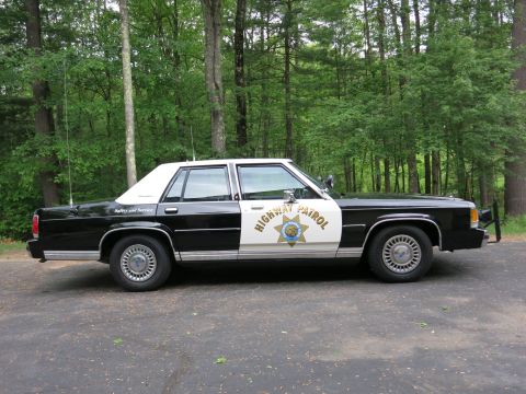 1991 Ford LTD Crown Victoria - Side view of a black-and-white Highway Patrol police sedan with gold star door emblem and twin antennas parked in front of dense green tree…