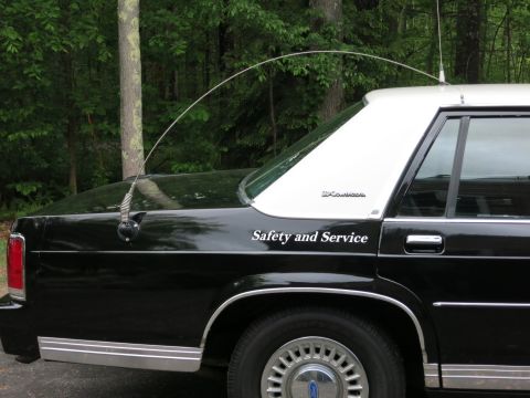 1991 Ford LTD Crown Victoria - Rear quarter of a black sedan with a curved whip antenna and white 'Safety and Service' lettering on the door, parked by trees.