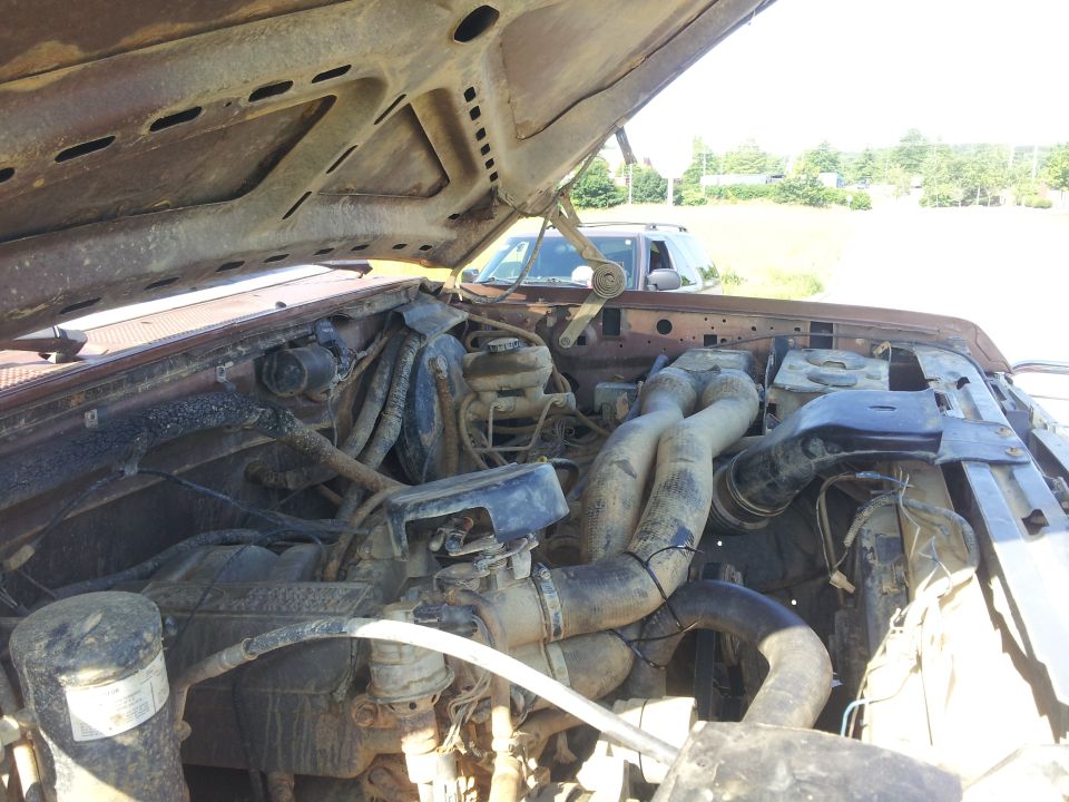 Rusty engine bay of a brown 1980s Ford truck with the hood open, showing hoses and engine components