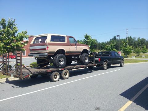 1988 Ford Bronco - A lifted Ford Bronco is transported on a trailer towed by a black Ford pickup truck under a clear blue sky.