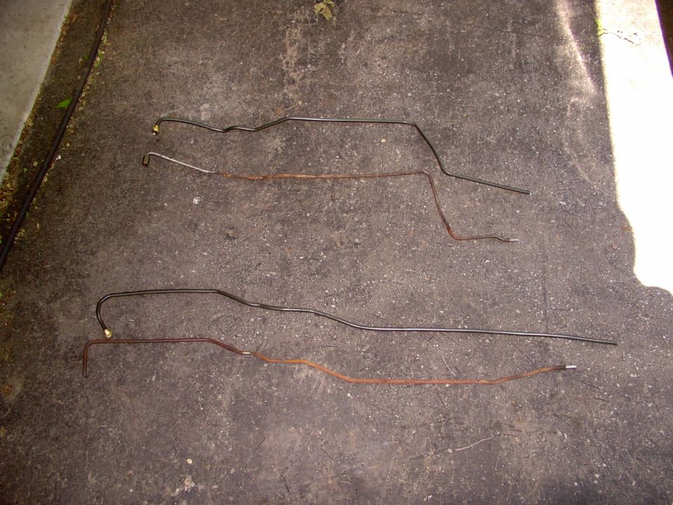 Four pre-bent metal fuel or brake lines, two new steel and two rusty originals, laid on concrete