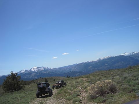 1989 Ford Bronco - Two off-road vehicles travel along a dirt path with snow-capped mountains in the distance under a clear blue sky.