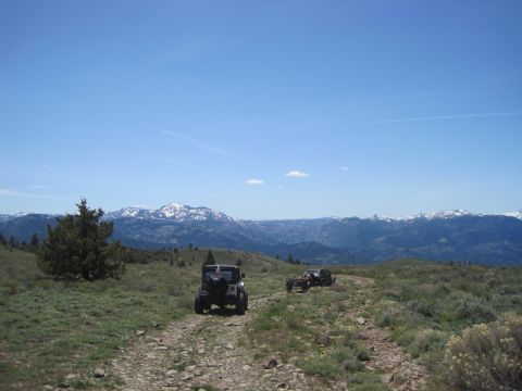 1989 Ford Bronco - Off-road vehicles navigate a mountain trail under a clear blue sky with distant snow-capped peaks.