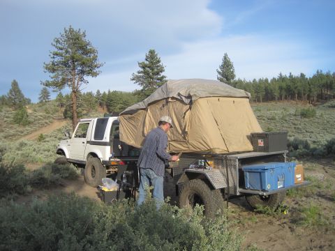 1989 Ford Bronco - A white SUV is equipped with a rooftop tent and a trailer, set up for camping in a dry, hilly landscape.