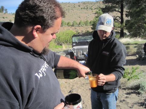 1989 Ford Bronco - Two men pour drinks in a sandy, outdoor setting with a Jeep in the background.
