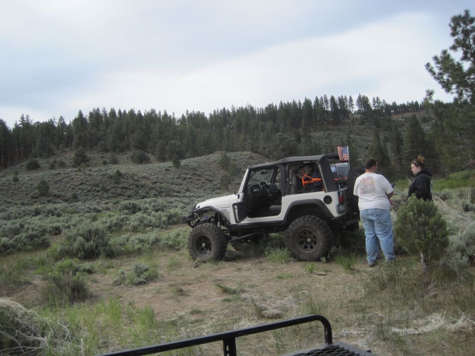 White Jeep Wrangler parked on a dirt path with two people standing nearby. American flag visible on the back