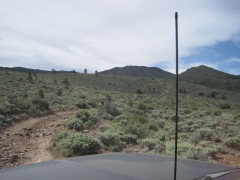 1989 Ford Bronco - Two off-road vehicles traverse a , dusty trail winding through a dry, scrub-covered hillside under a partly cloudy sky.