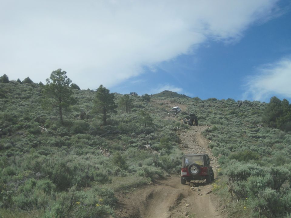 Several off-road vehicles driving up a steep, rocky dirt path on a scrub-covered hillside under a partly cloudy sky