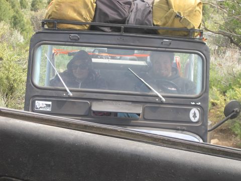 1989 Ford Bronco - Two people are visible through the windshield of a Jeep CJ-7, which has luggage strapped to its roof rack.