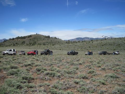 1989 Ford Bronco - A line of off-road vehicles is parked in a vast, open field with rolling hills and distant snow-capped mountains under a bright blue sky.
