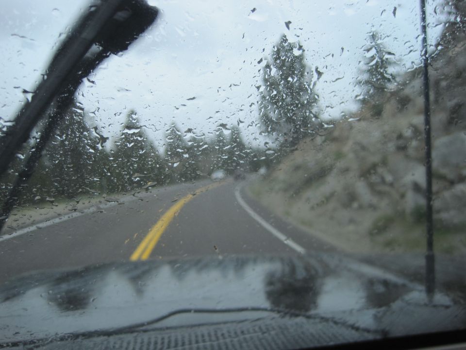 View from inside a vehicle driving on a road with rain on the windshield. Trees and a rocky hillside are visible
