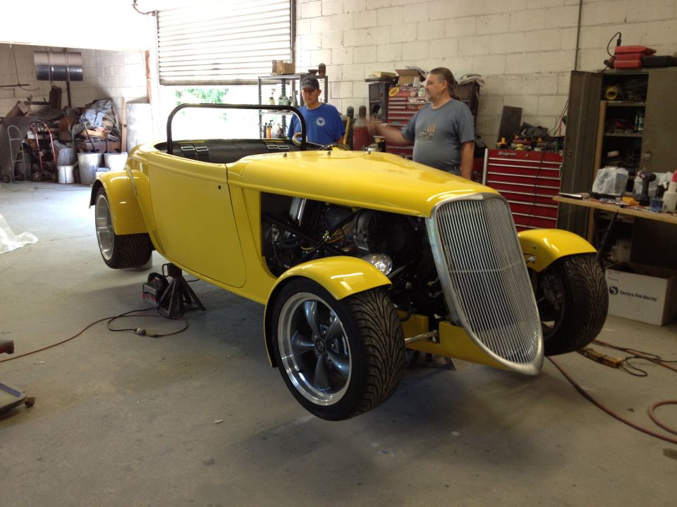 Yellow hot rod roadster with exposed engine and large chrome grille, being worked on in a garage