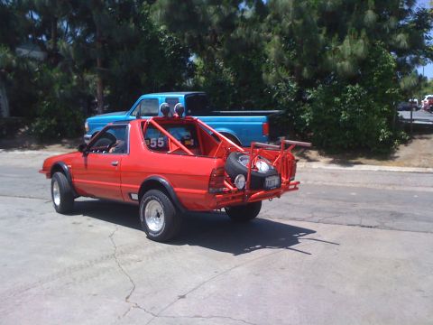 1968 Ford Mustang - A modified red Subaru BRAT is parked behind a blue pickup truck, off-road modifications and a spare tire carrier.