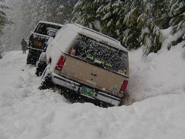 Snow-covered tan SUV stuck in deep snow on a wooded road, rear 3/4 view