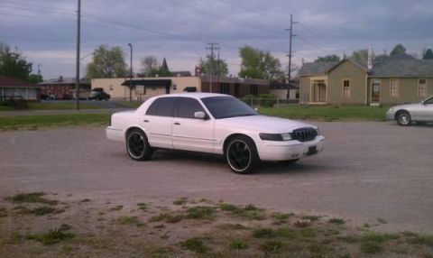 2000 Mercury Grand Marquis - A white 2000 Mercury Grand Marquis sedan is parked on a gravel lot with buildings in the background.