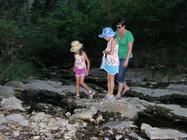 Woman and two girls wearing hats walk across rocks in a shallow stream bed