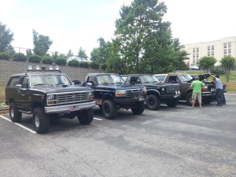 1986 Ford Bronco - A line of Ford Broncos and trucks are parked in a lot, with a few people gathered around one of the vehicles.
