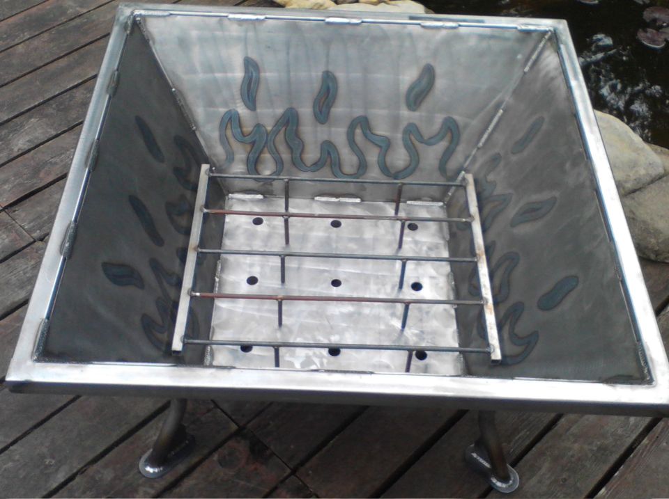 Square metal fire pit with grate inside, sitting on wooden deck. Flame shapes cut into the inner walls