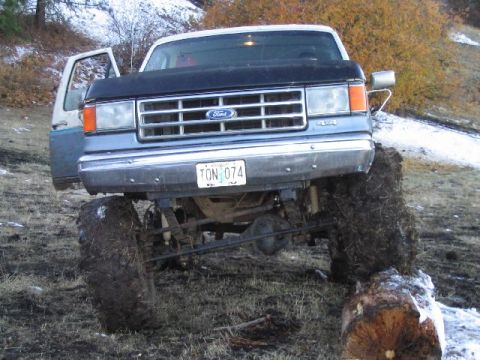 1988 Ford Bronco - A lifted Ford Bronco with mud-caked tires is positioned on uneven terrain with its driver's door ajar.