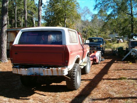 1979 Ford Bronco - A red Ford Bronco is loaded onto a trailer, with a black truck hitched to it, in a wooded area.