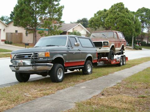 1979 Ford Bronco - A gray Ford Bronco tows a red Ford Bronco on a trailer down a suburban street.