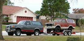 1979 Ford Bronco - A gray 1979 Ford Bronco tows a red Bronco on a trailer down a suburban street.