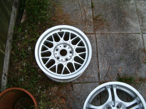 1991 Ford Bronco - A close-up view of two silver alloy wheels, one a detailed mesh pattern, placed outdoors on a paved surface.
