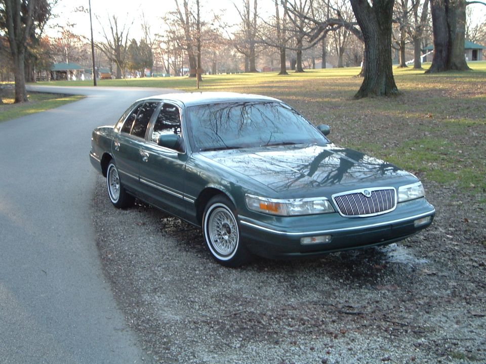 Green Mercury Grand Marquis sedan with whitewall tires parked on a road in a wooded park