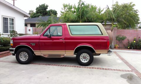 1994 Ford Bronco - Driver's-side profile of a red and tan two-tone full-size Ford Bronco 4x4 parked on a concrete driveway with palm trees and a block wall be…