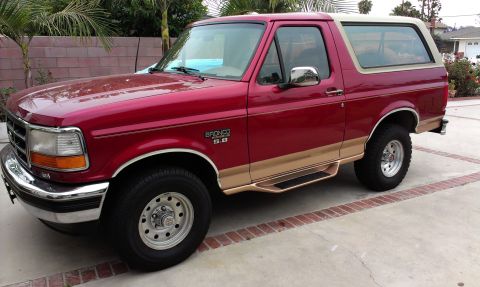 1994 Ford Bronco - Driver's-side front three-quarter view of a red and tan two-tone full-size Ford Bronco 4x4 on a concrete driveway, backed by a block wall a…