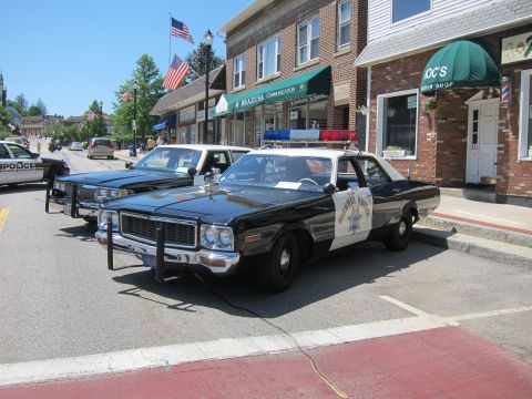 1991 Ford LTD Crown Victoria - Two black-and-white police cruisers, one a vintage Highway Patrol sedan with light bar, parked along a brick downtown street with shops.