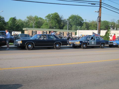 1991 Ford LTD Crown Victoria - row of vintage police cars across a street, including a black sedan and a State Police cruiser, with motorcycles parked behind them.