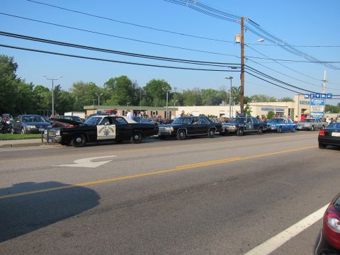 1991 Ford LTD Crown Victoria - row of classic police cruisers, led by a black-and-white Highway Patrol sedan, parked along a street with crowds and shops in the backgroun…