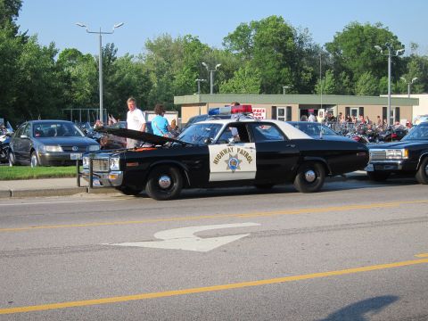 1991 Ford LTD Crown Victoria - black-and-white Highway Patrol sedan with roof light bar and chrome grille parked along a road at a crowded outdoor event with motorcycles.