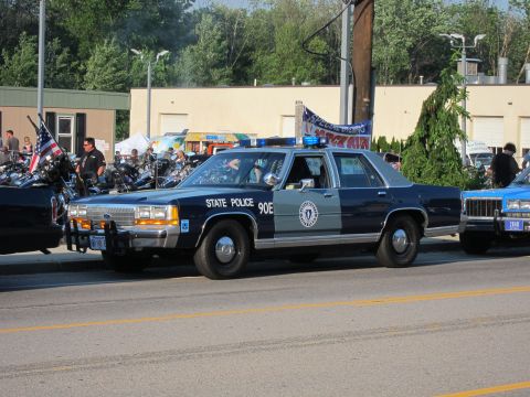 1991 Ford LTD Crown Victoria - dark-blue and silver Ford State Police sedan marked '90E' with a state seal and roof light bar, parked at a crowded roadside event.