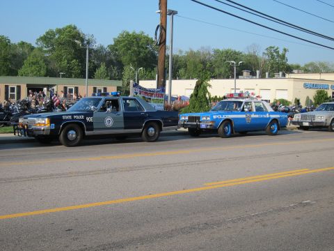 1991 Ford LTD Crown Victoria - dark-blue State Police sedan marked 90E beside a light-blue marked Police cruiser, parked along a street with motorcycles behind.