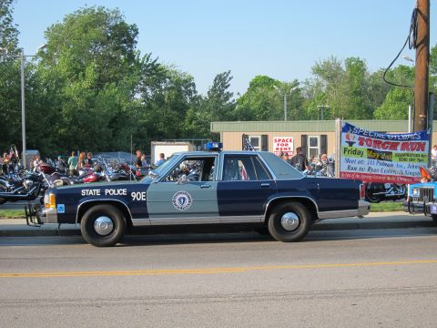 1991 Ford LTD Crown Victoria - dark-blue and silver Ford State Police sedan marked '90E' with roof light bar, parked along a road by a Special Olympics Torch Run banner.