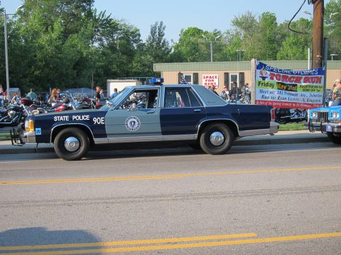 1991 Ford LTD Crown Victoria - Side view of a dark-blue and silver Ford State Police sedan marked '90E' parked along a road near a Torch Run banner and motorcycles.
