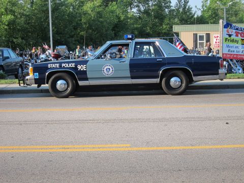1991 Ford LTD Crown Victoria - Side profile of a dark-blue and silver Ford State Police cruiser marked '90E' with roof light bar, parked along a street with bikes behind.